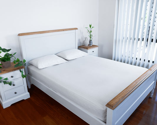 A neatly made bed with white grounding sheets in a bright, minimalist bedroom featuring matching white furniture with wooden accents, potted plants, and vertical blinds letting in natural light.